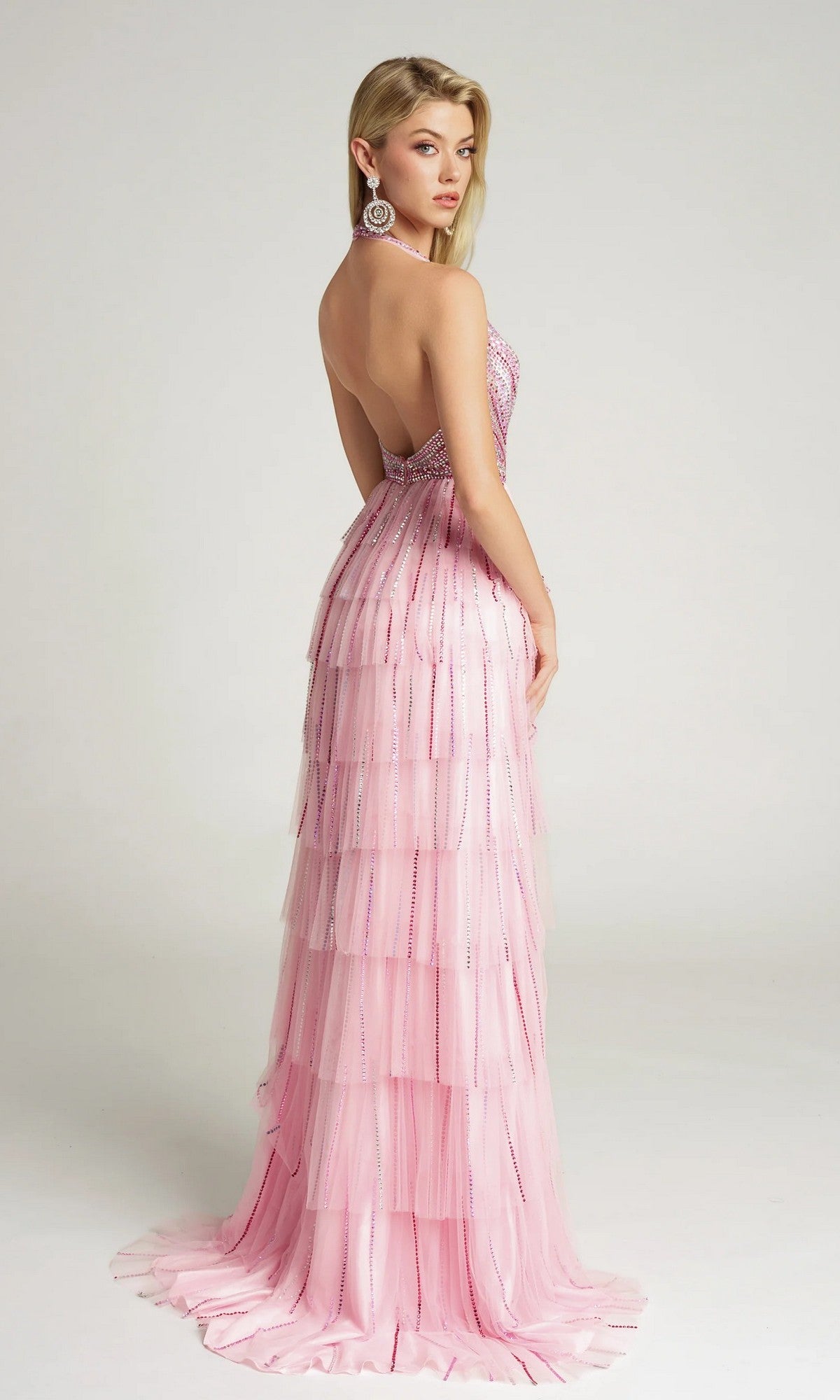 A woman poses in a studio wearing the Portia and Scarlett PS26117 long formal dress, a pink strapless, backless gown with layered ruffles and sparkling beadwork, looking over her shoulder.